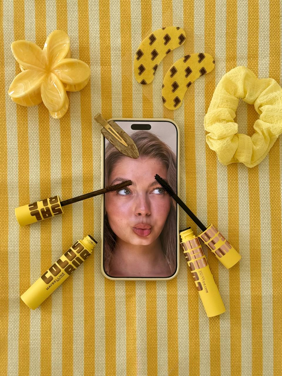 Maybelline mascara with a phone portrait and hair accessories on yellow striped fabric